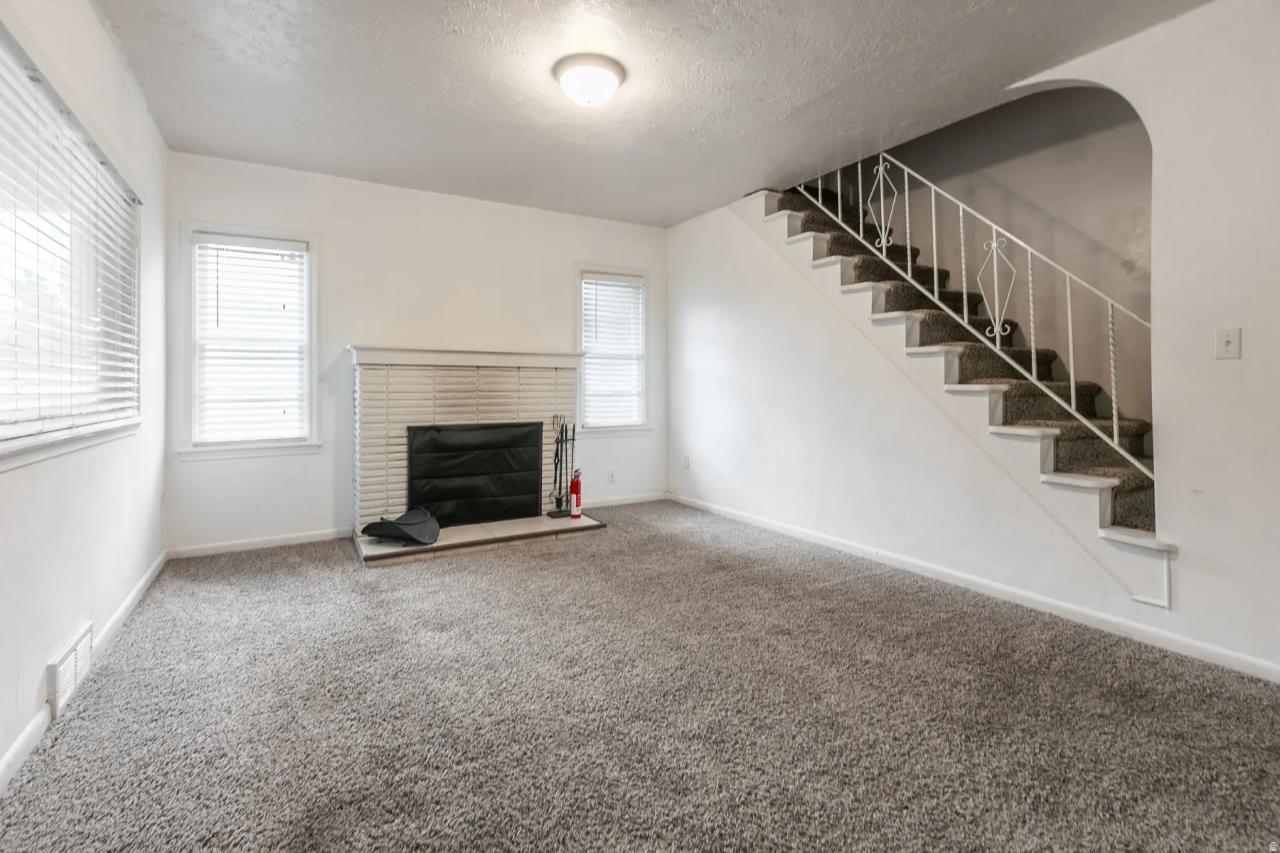 Spacious room featuring a white brick fireplace with a black insert, light grey carpeting, multiple windows with blinds, and a staircase with white wrought iron railing