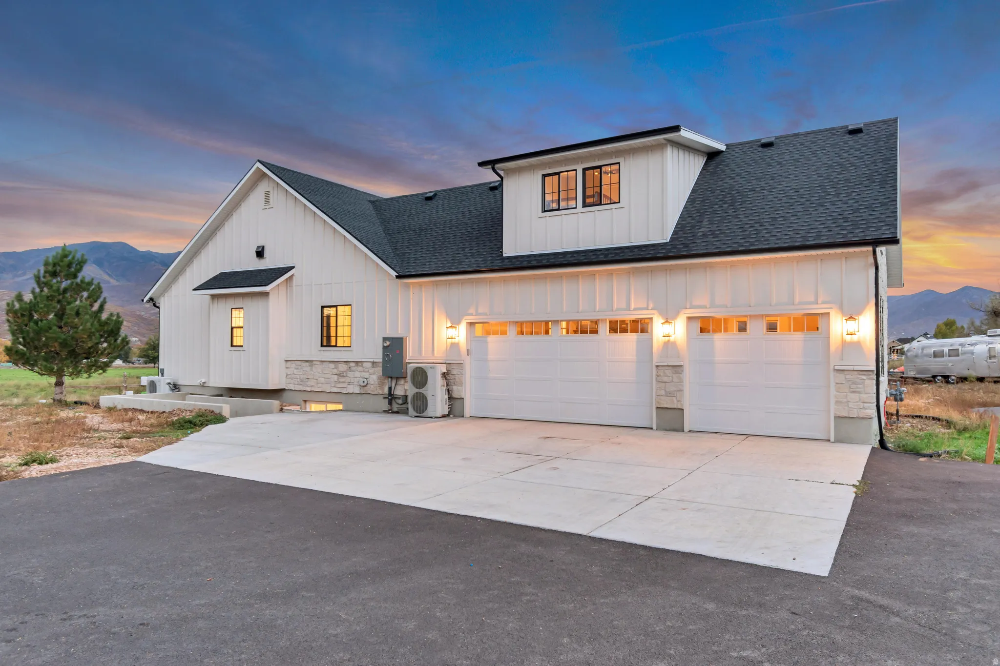 Modern farmhouse exterior featuring board and batten siding, stone accents, a dark shingle roof, a two-car garage with transom windows, and a paved driveway
