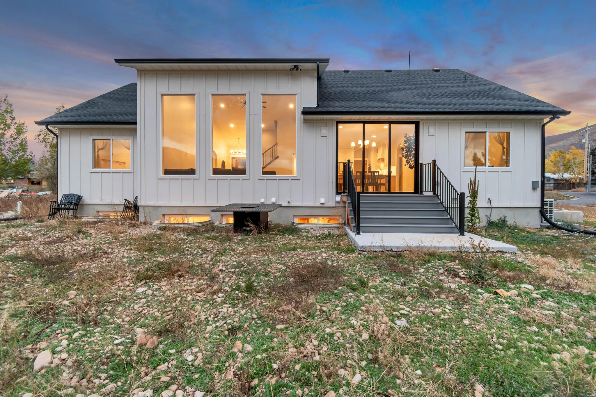 Contemporary exterior featuring white board and batten siding, multiple large windows, a dark shingle roof, and a rear patio with stairs