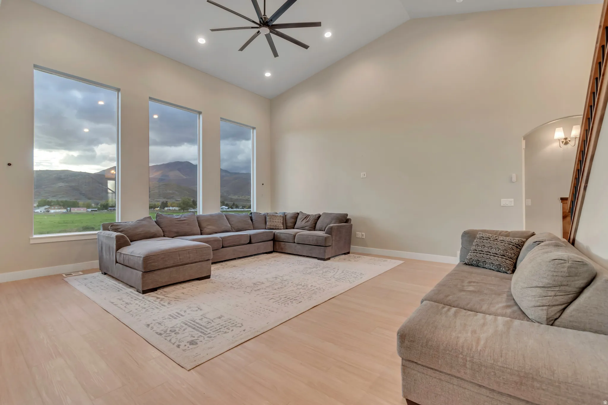 Living area featuring vaulted ceilings, three tall windows with mountain scenery, light wood-finish flooring, recessed lighting, and a modern ceiling fan