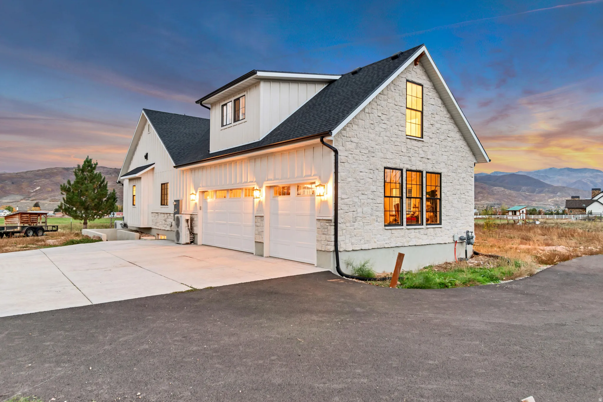 Modern farmhouse exterior featuring vertical white board and batten siding, light-toned stacked stone accents, a dark shingled roof, a two-car garage with carriage-style doors, and black-framed windows