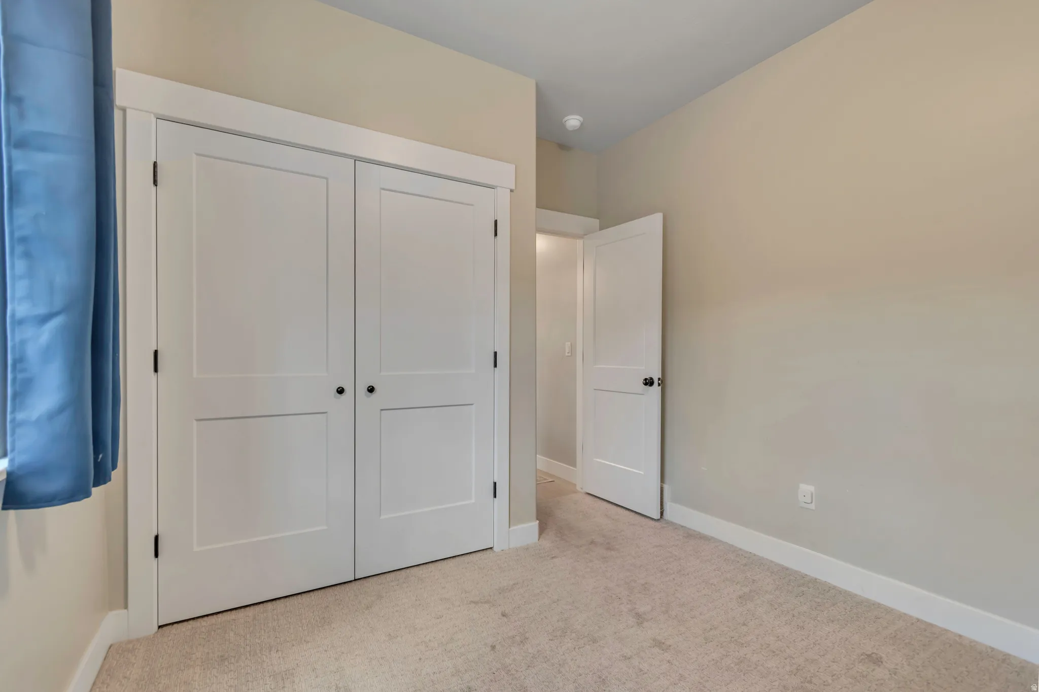 Neutral-toned room featuring a double-door closet with modern black hardware, a single panel interior door, neutral carpeting, and white baseboards