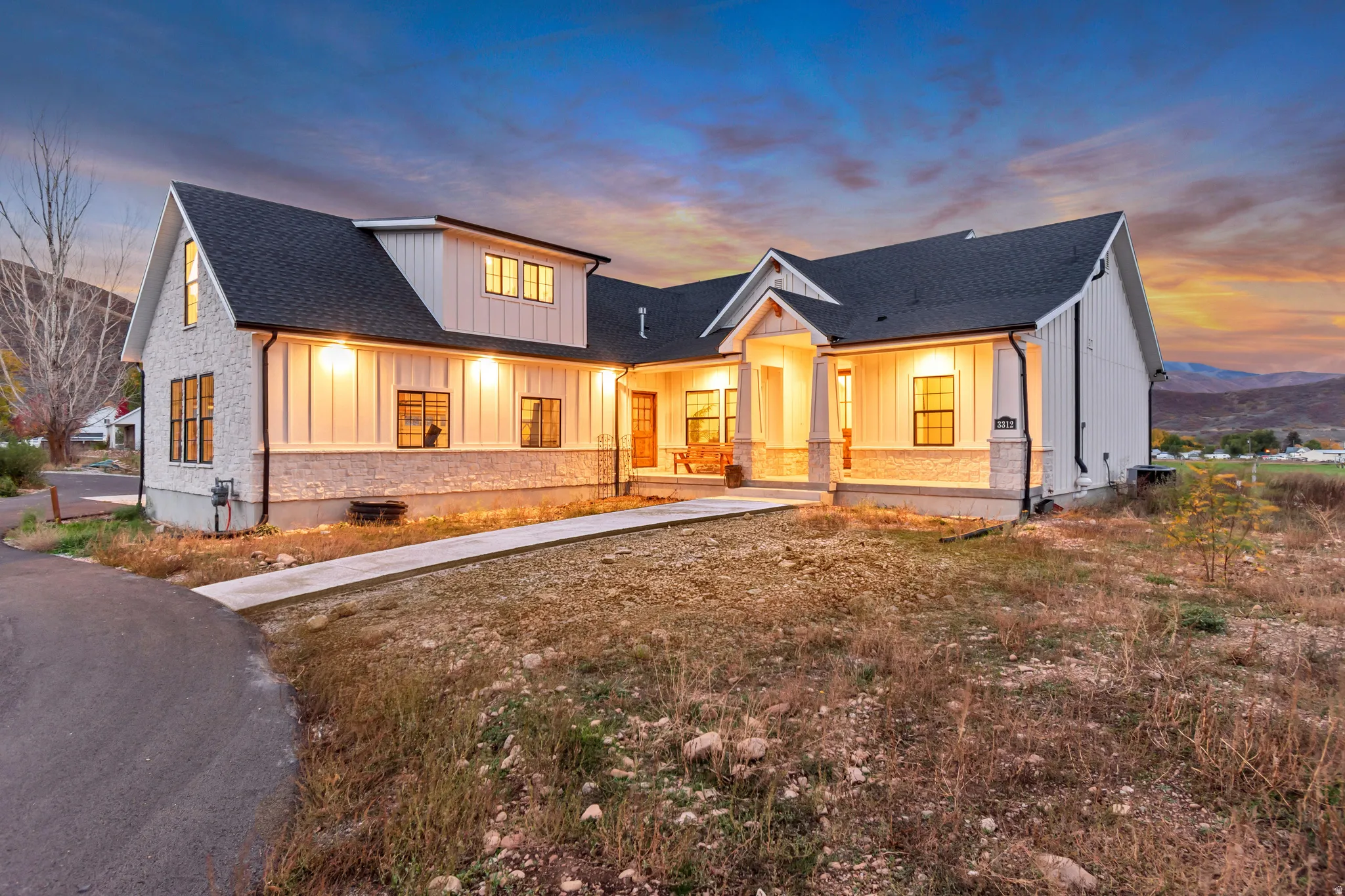 Modern farmhouse exterior with white board-and-batten siding, light-colored stone accents, a dark asphalt shingle roof, black-framed windows, and an inviting covered porch