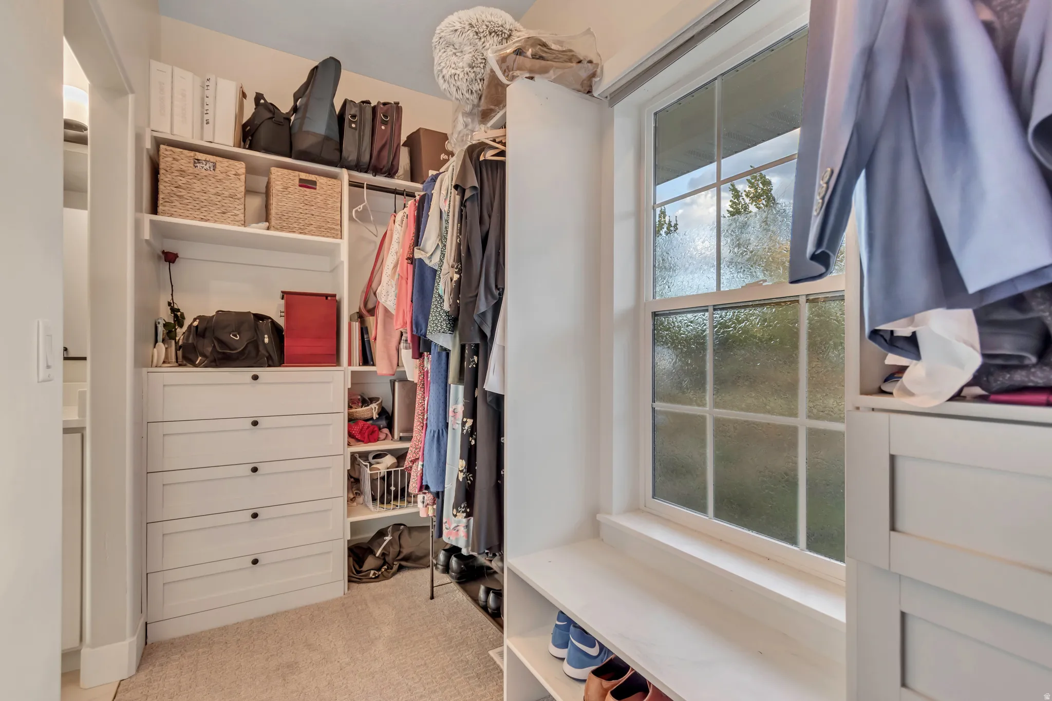 Built-in white custom closet system featuring multiple shelving units, hanging rods, an integrated five-drawer dresser, and a window with a built-in bench