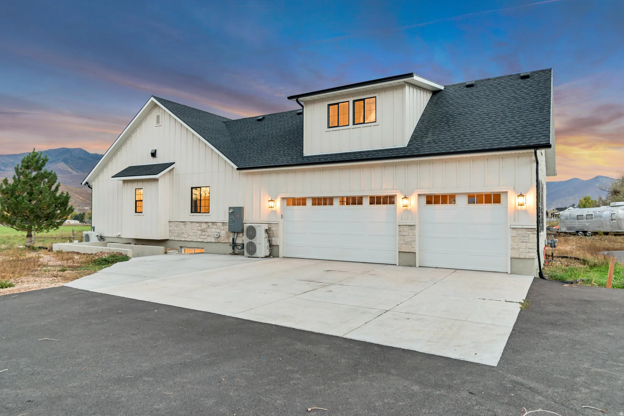 Modern farmhouse exterior featuring white board-and-batten siding, a black asphalt shingle roof, a two-car garage with transom windows, stone accents, and exterior wall sconces