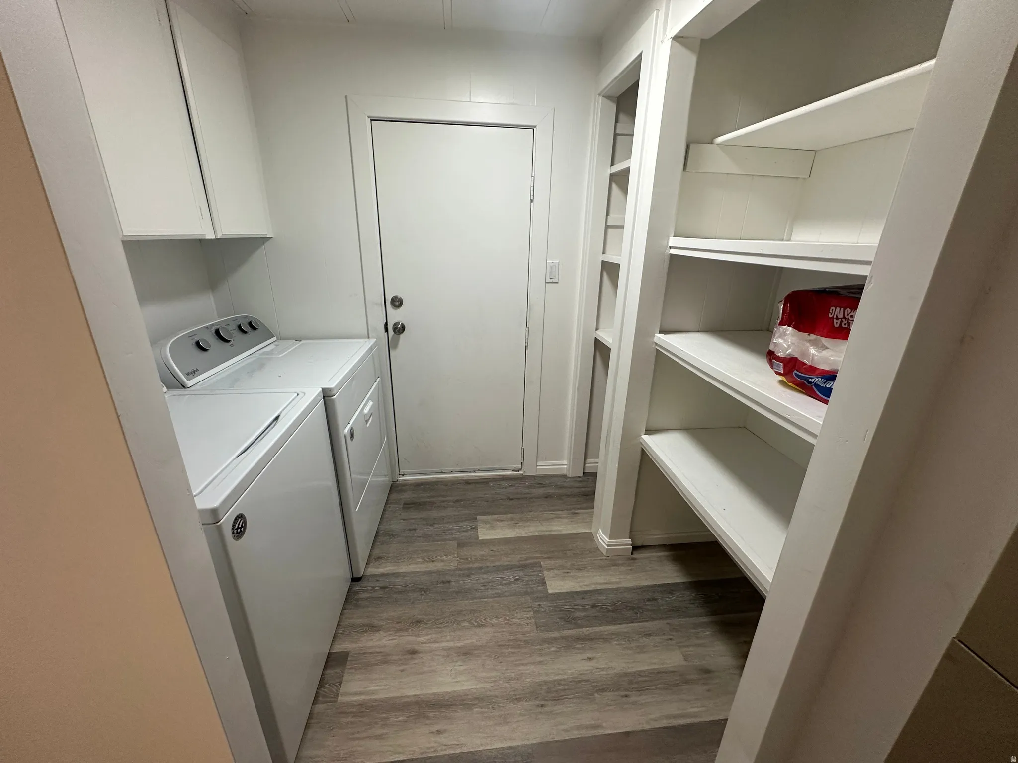 Laundry area featuring wood-finish flooring, white built-in shelving, white overhead cabinetry, and a solid white door