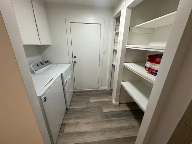 Laundry area featuring wood-finish flooring, white built-in shelving, white overhead cabinetry, and a solid white door