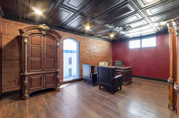 Spacious room featuring exposed brick walls, wood-finish flooring, a decorative coffered ceiling, and an arched window