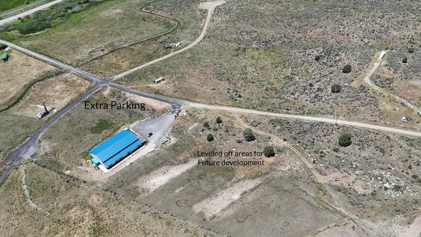 Aerial perspective of a property featuring a building with a blue metal roof, an expansive gravel parking area, and unpaved access roads