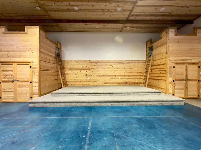 Open-concept space featuring a raised stage with carpeted steps, natural wood paneling, wood-finish ceiling, exposed wood beams, and a blue-painted floor