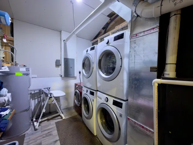 Laundry area featuring wood-finish flooring, two stacked front-loading washer and dryer units, a water heater, and exposed overhead piping