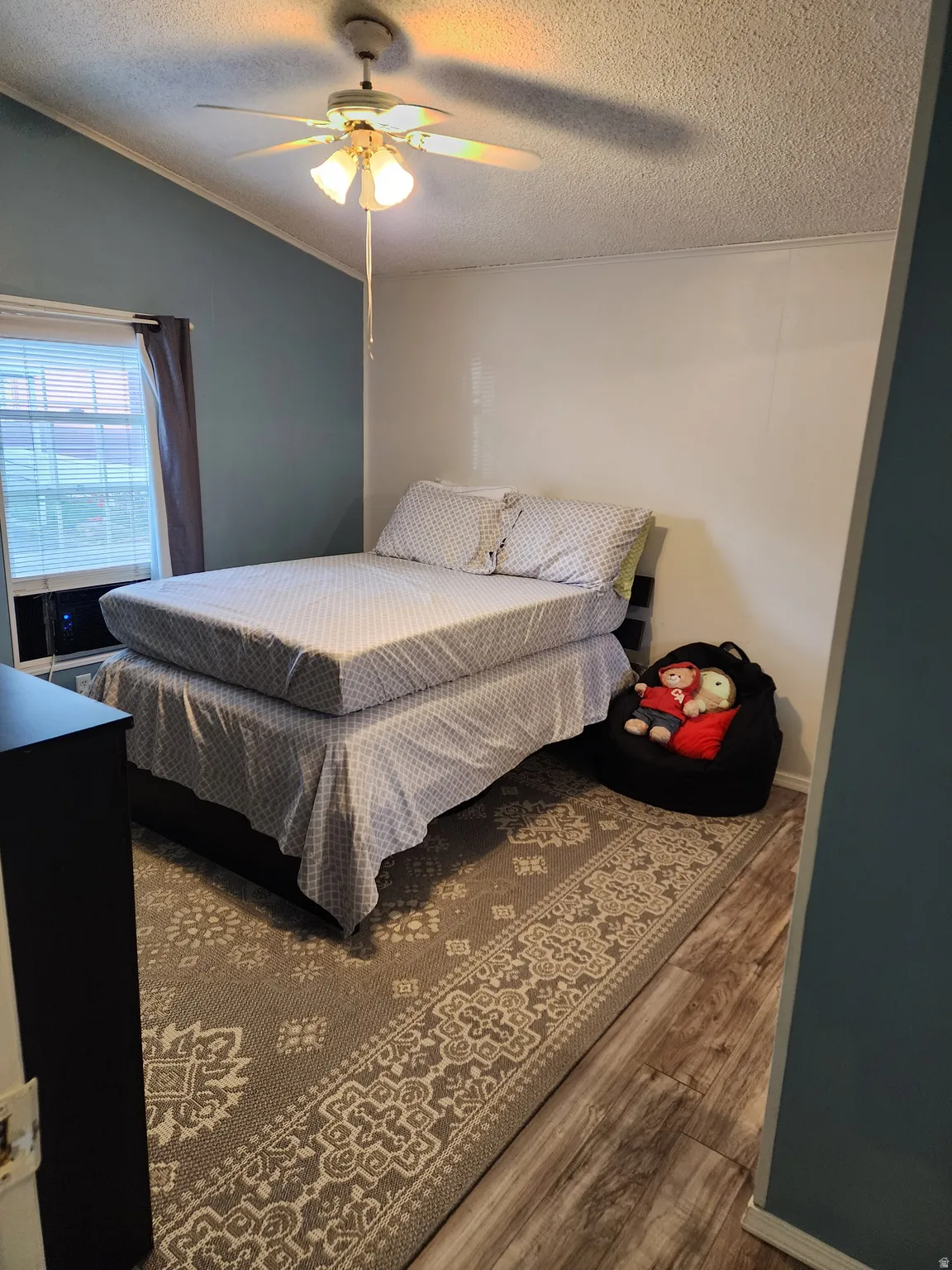 Spacious room featuring vaulted ceilings, a ceiling fan with integrated lighting, wood-finish flooring, and a large window with blinds