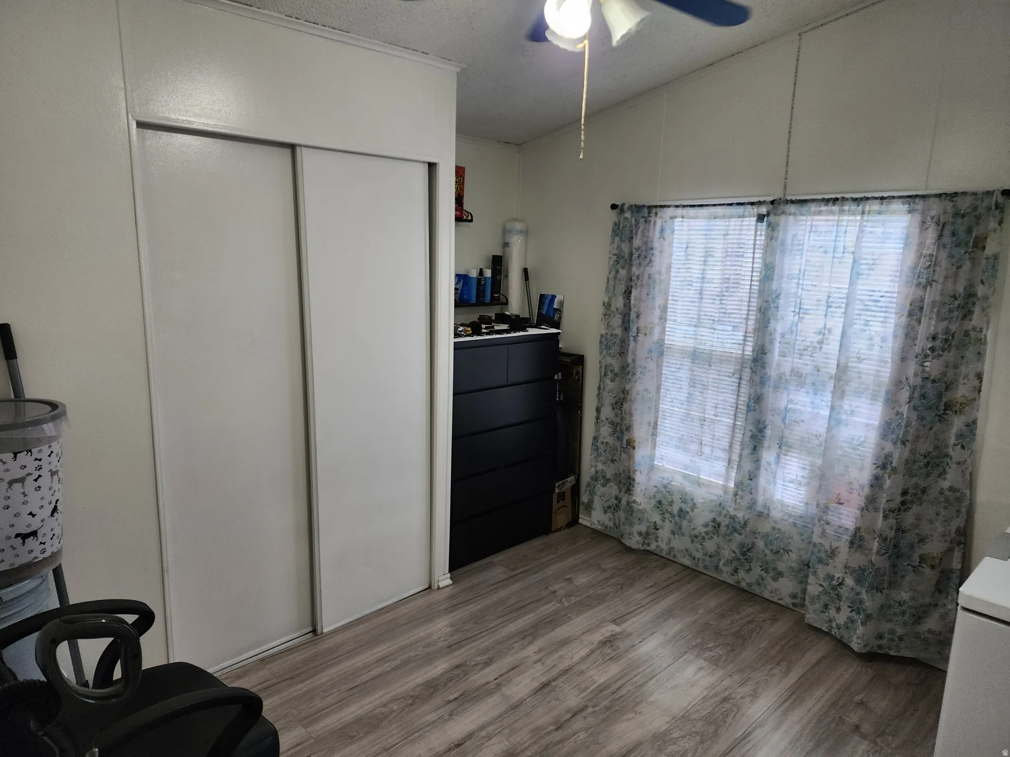 Interior room featuring wood-finish flooring, white paneled walls, and a window with blinds and floral pattern curtains