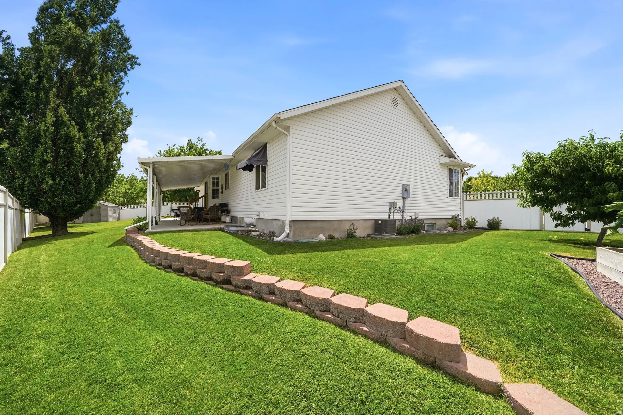Expansive rear yard featuring a covered patio, a decorative retaining wall, white vinyl fencing, mature trees, and a detached storage shed