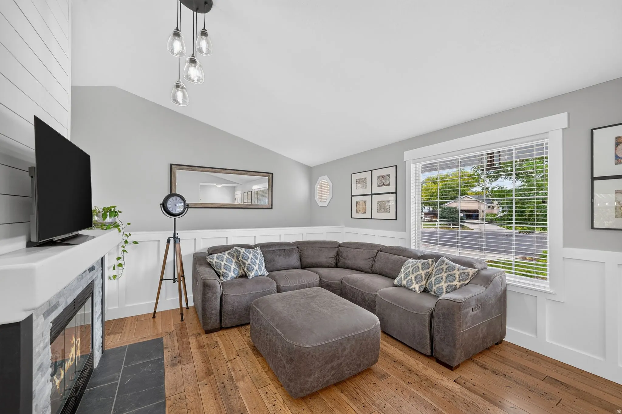 Living area featuring hardwood flooring, a fireplace with a stone surround and hearth, wainscoting, and a vaulted ceiling with a contemporary pendant light fixture