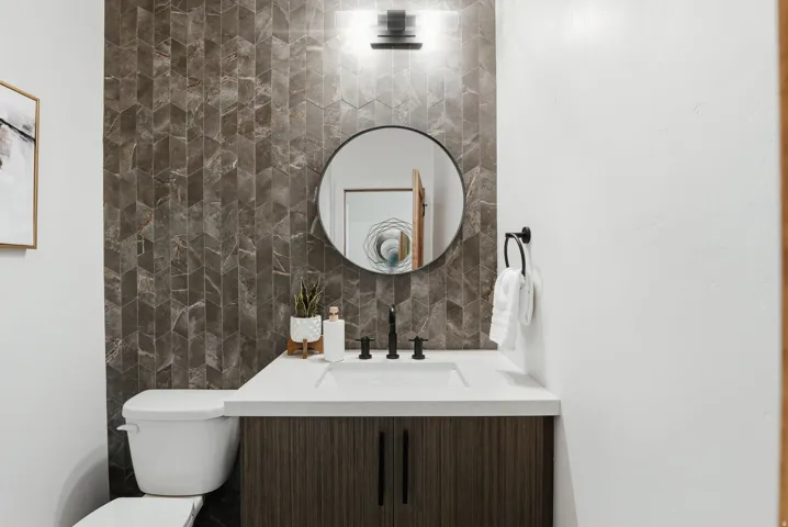 Main floor half bath, vanity with multi-patterned tile accents, white countertop, integrated sink, and wood-finish cabinetry