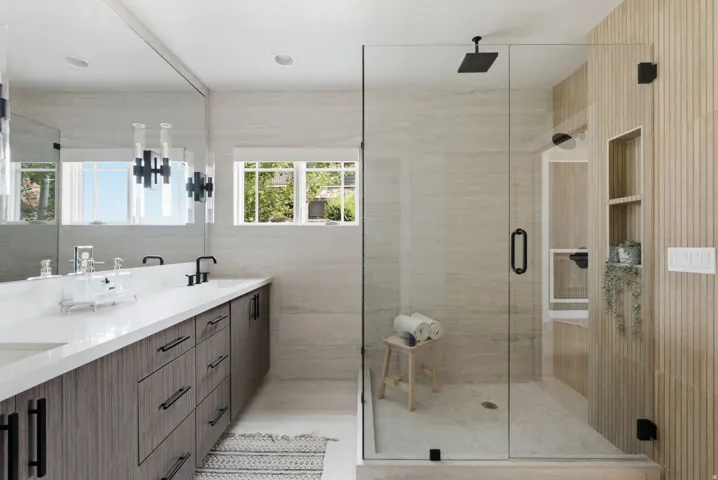 Modern bathroom featuring a glass-enclosed shower with a matte black rain showerhead, a dual-sink vanity with a white countertop and wood-finish cabinetry, and light-toned wall and floor tile