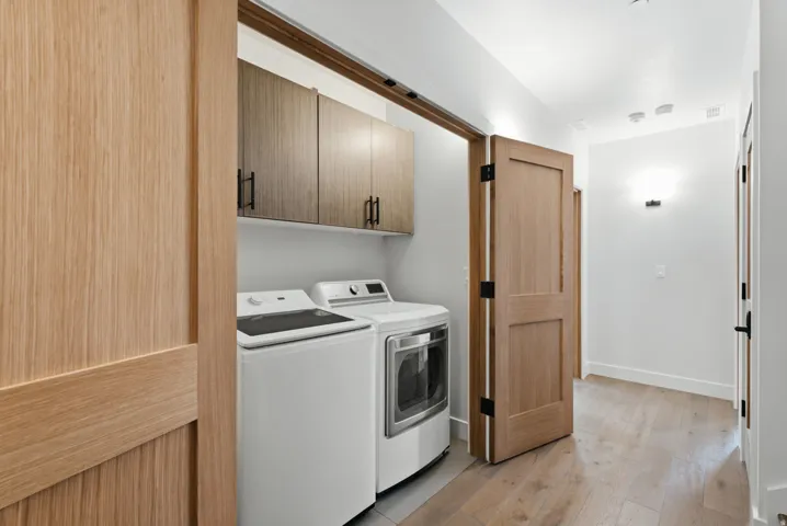 Laundry area w/ washer & dryer, wood-finish flooring, built-in cabinetry with matte black hardware, and an open-concept hall with recessed lighting