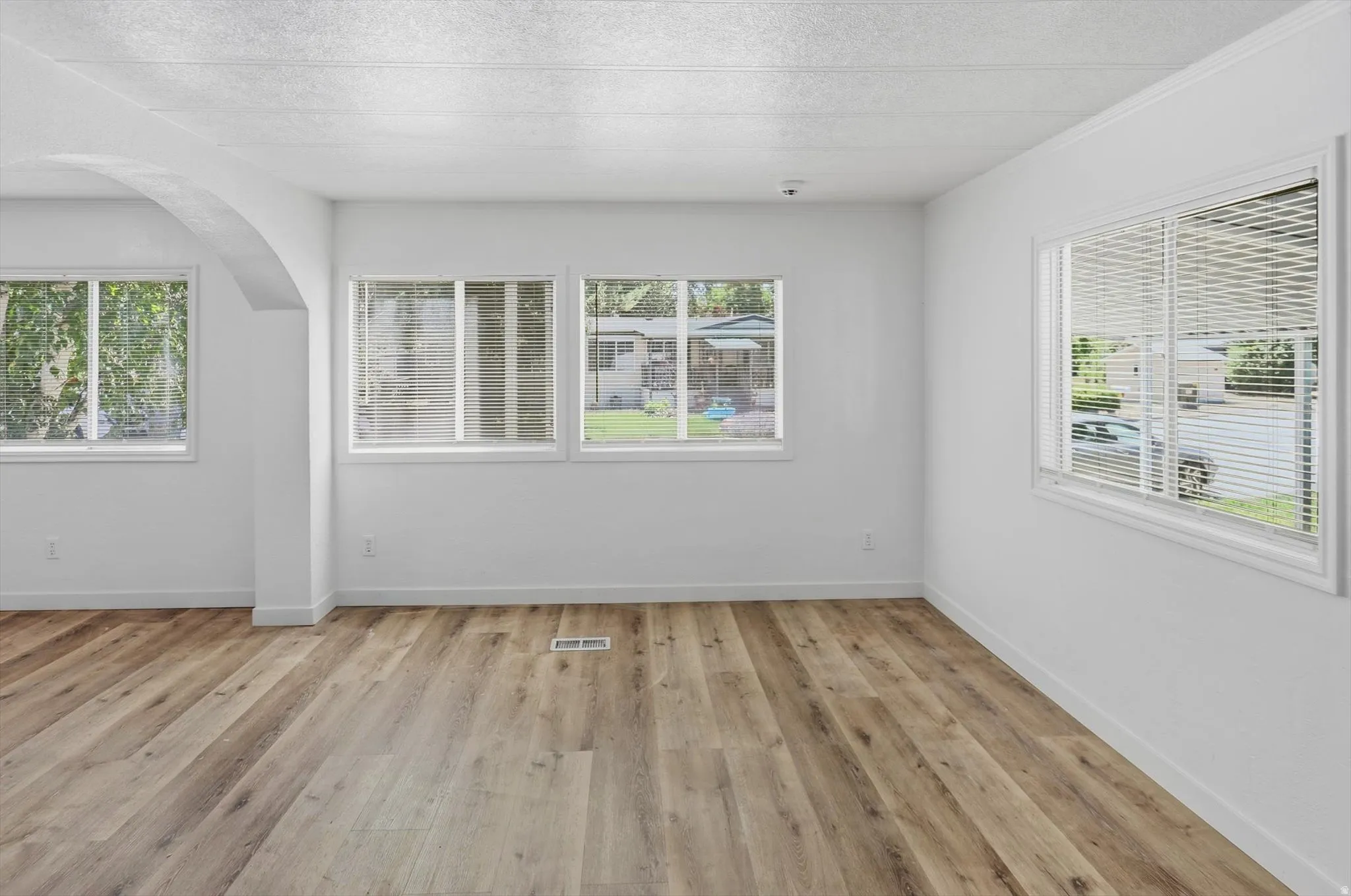 Spacious room featuring wood-finish flooring, white walls, multiple windows with blinds, and an arched entryway