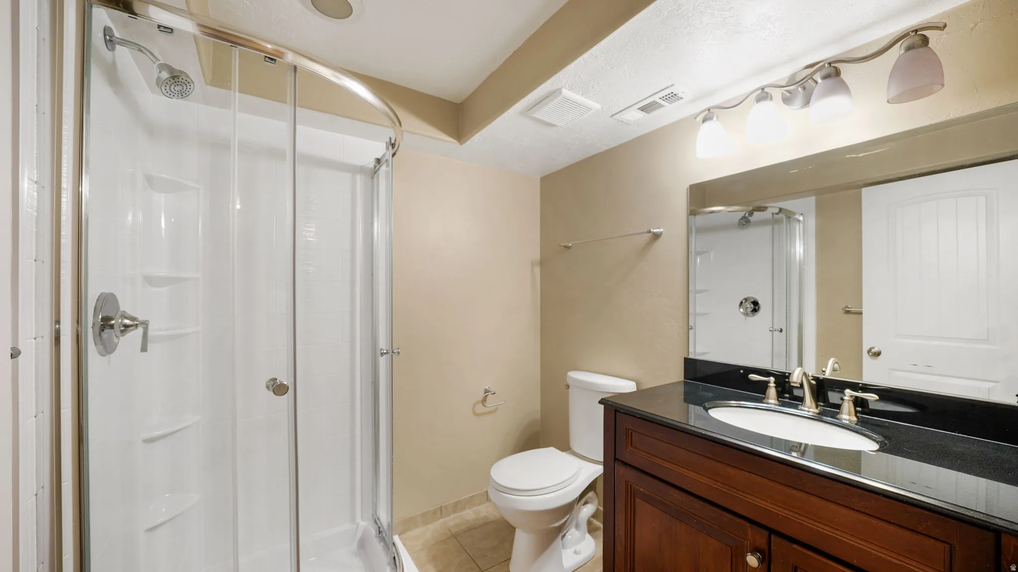 Bathroom featuring a white shower enclosure with a curved glass door, a dark wood-finish vanity with a dark countertop and integrated sink, a large wall-mounted mirror, and tile flooring