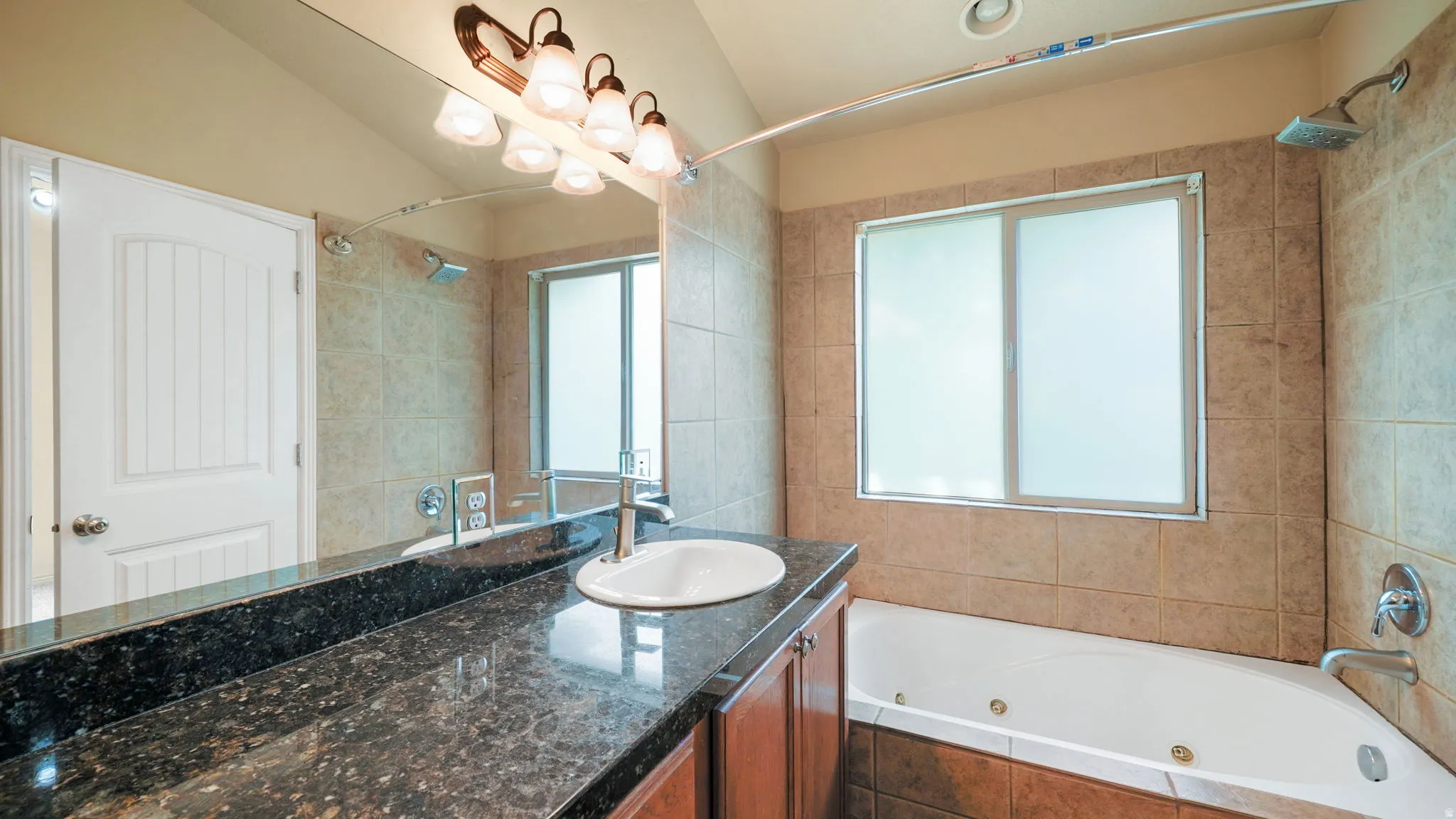 Bathroom featuring a built-in jetted tub, tile surround, and a vanity with a dark speckled countertop and undermount sink