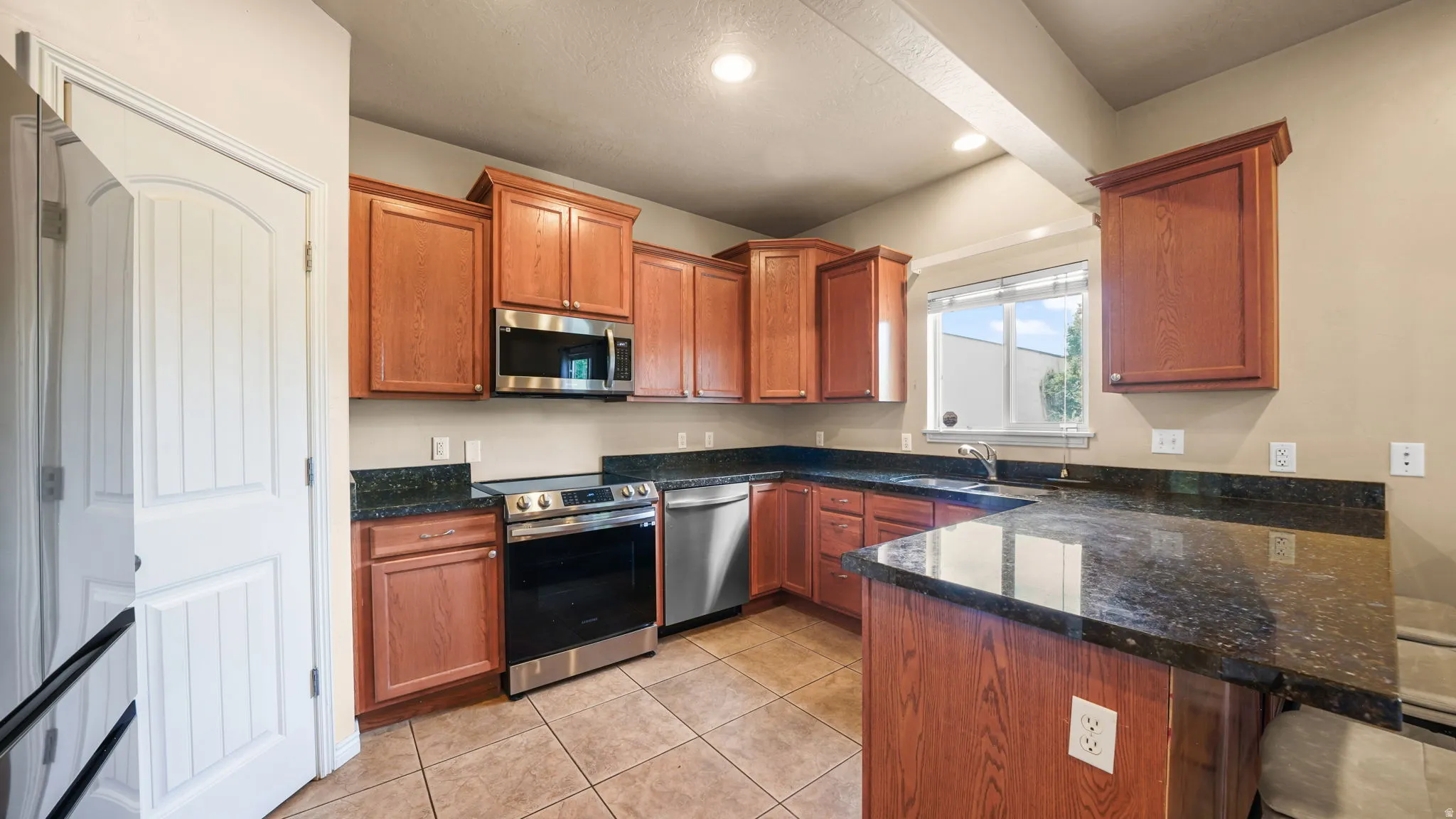Kitchen featuring wood cabinetry, dark countertops, tile flooring, stainless steel appliances, and an undermount sink