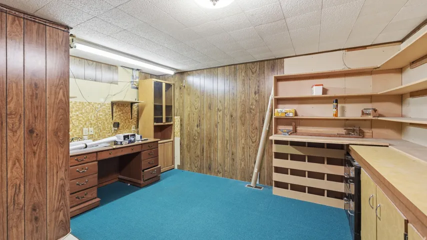 Room with wood paneling and teal carpeting, featuring built-in shelving, a built-in workbench with storage, fluorescent lighting, and a drop ceiling