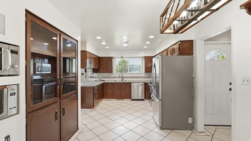 Bright kitchen featuring wood cabinetry, white tile flooring, stainless steel appliances, recessed lighting, and a decorative overhead light fixture