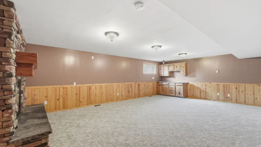 Spacious room featuring a stone-clad fireplace with a stone hearth, wood-finish wainscoting, light-colored carpeting, and a built-in cabinet and counter area with upper cabinetry