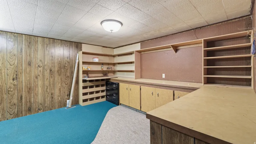 Room featuring wood-look paneling, built-in shelving, built-in cabinetry, pegboard wall, and a drop ceiling with a flush-mount light fixture