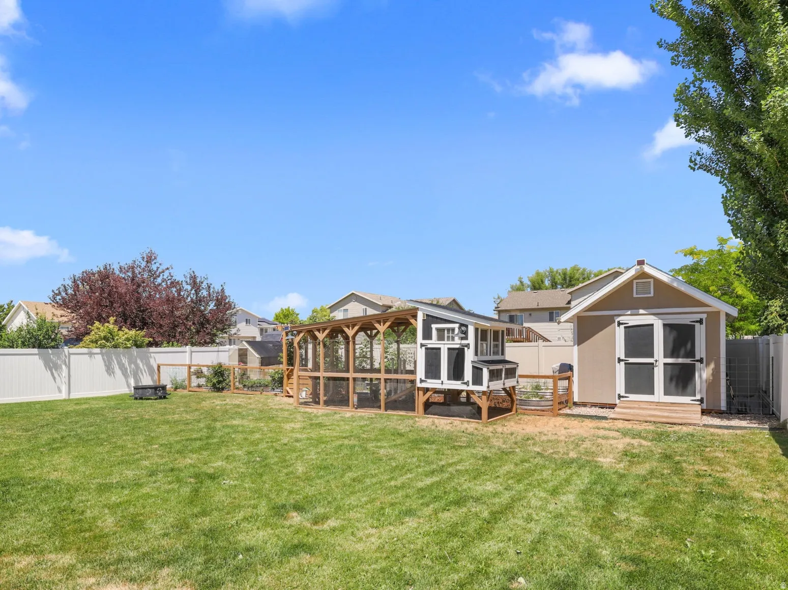 Expansive green lawn featuring a substantial storage shed with double doors, an enclosed chicken coop structure, and a perimeter white vinyl fence
