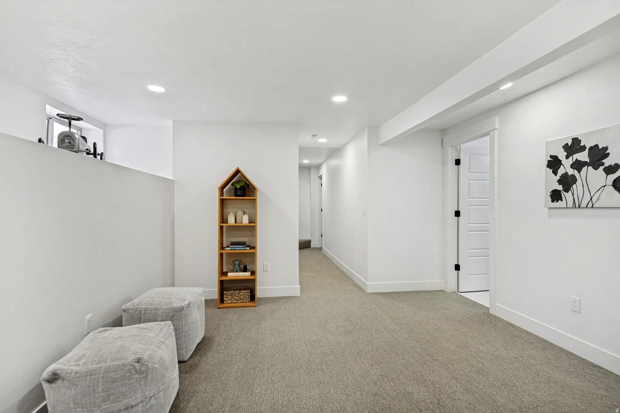 Bright, open room featuring recessed lighting, a neutral carpeted floor, white walls, and a visible doorway with a contrasting dark hardware