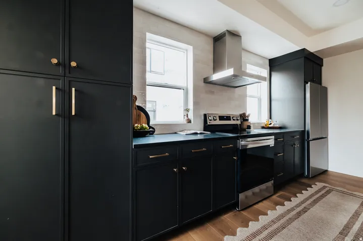 Modern kitchen featuring dark cabinetry with gold-tone hardware, stainless steel appliances, a tile backsplash, and wood-finish flooring