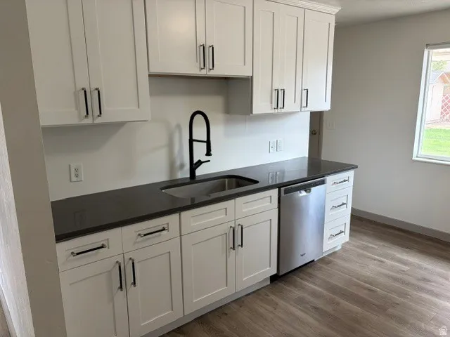 White shaker-style cabinetry with matte black hardware, dark solid surface countertops, stainless steel dishwasher, and wood-finish flooring