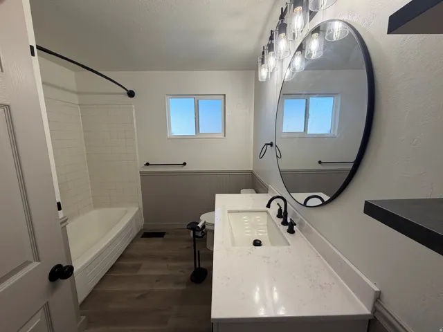 Bathroom featuring a white subway tile shower surround, wood-finish flooring, a vanity with a white countertop and rectangular undermount sink, and a large oval mirror with an overhead light fixture
