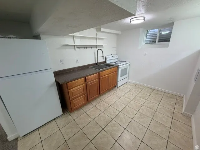 Full-size white refrigerator, white electric range, wood-finish cabinetry, dark countertop, and tile flooring