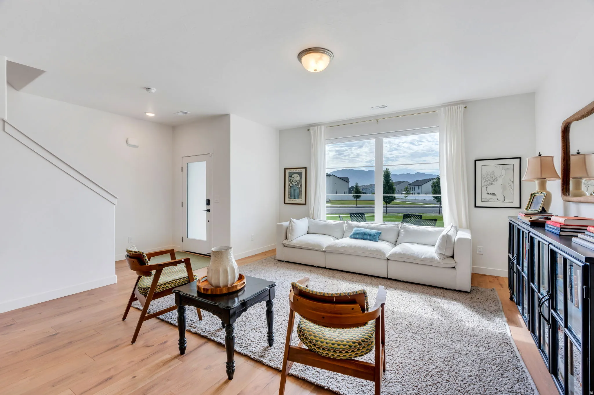 Spacious living area featuring expansive windows, wood-finish flooring, a contemporary overhead light fixture, a staircase with metal railings, and a glass-paneled entry door