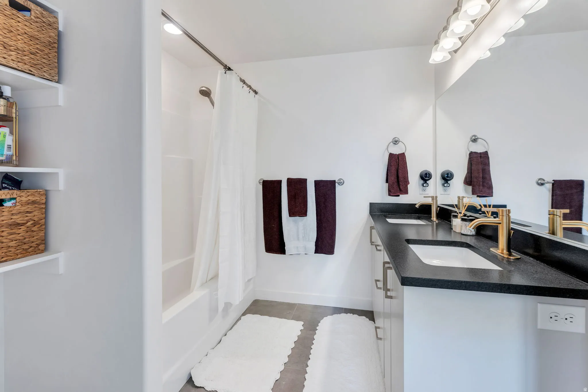 Bathroom featuring a dual vanity with a dark countertop and gold-finish fixtures, large mirror, and overhead light bar