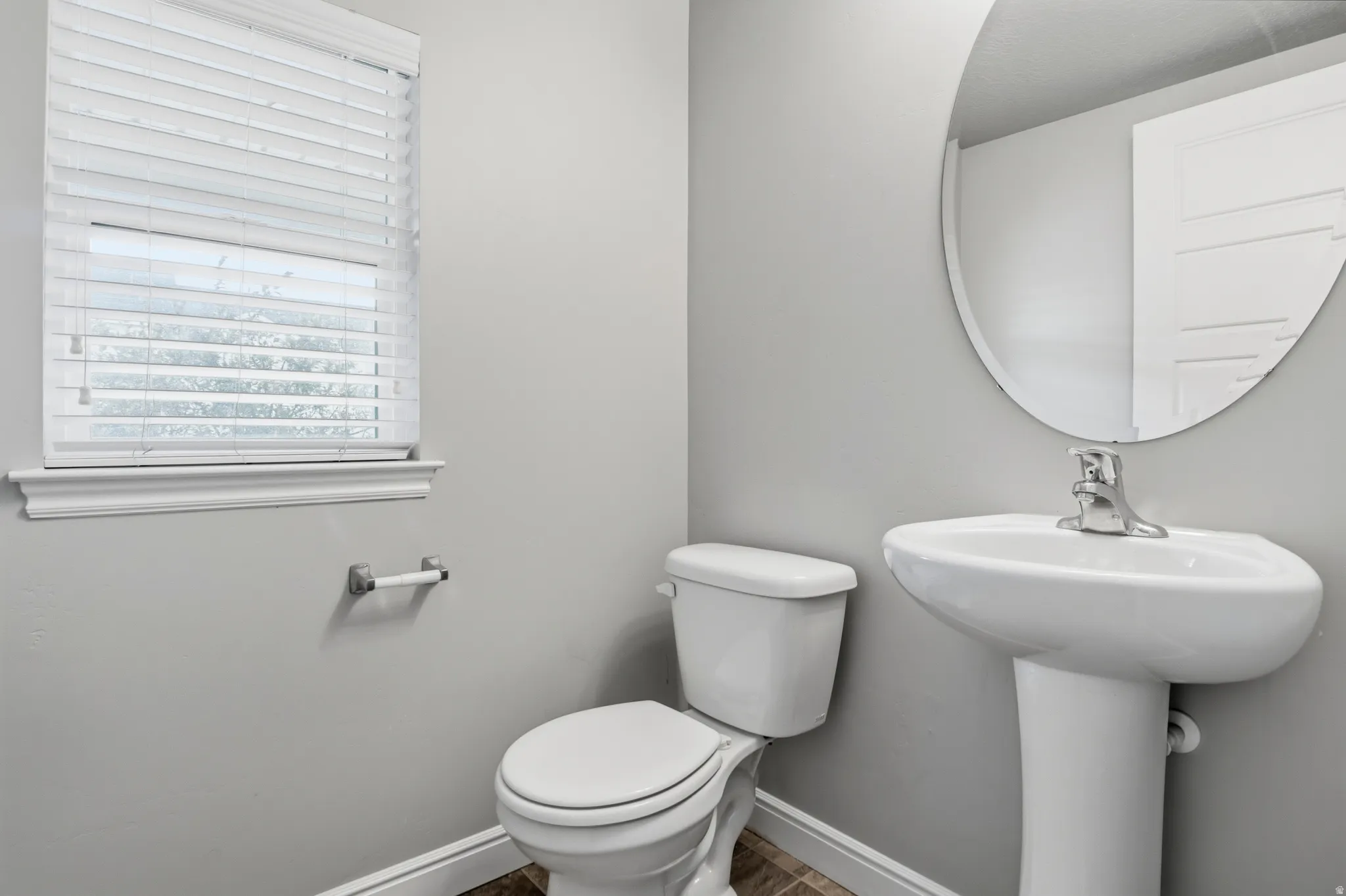 Pedestal sink with chrome faucet, oval frameless mirror, and a window featuring horizontal blinds