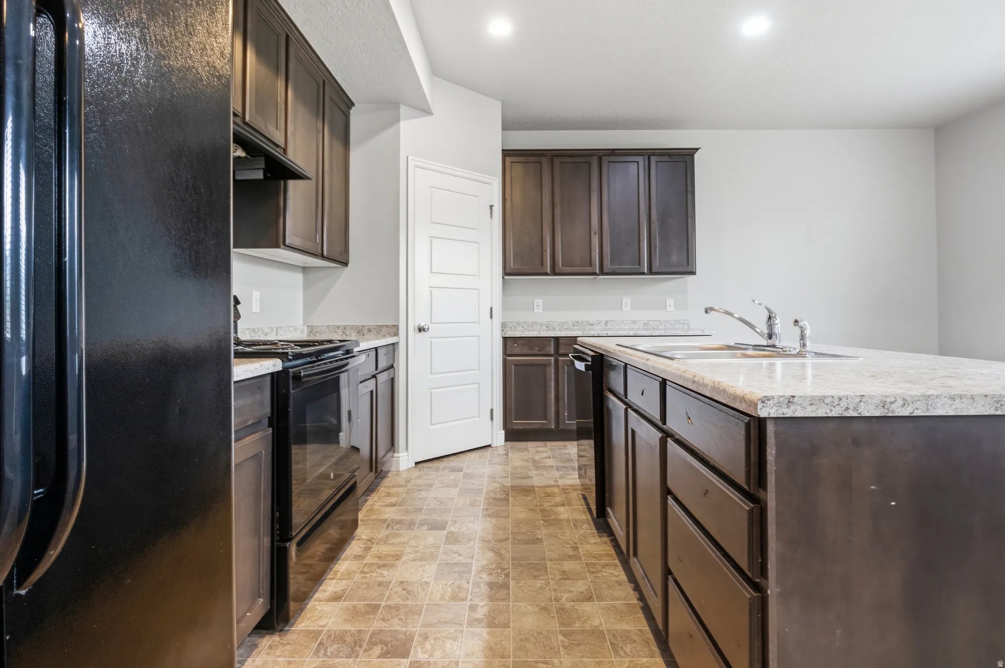 Kitchen featuring dark wood-finish cabinetry, light speckled countertops, a center island with an undermount sink, black appliances, and recessed lighting