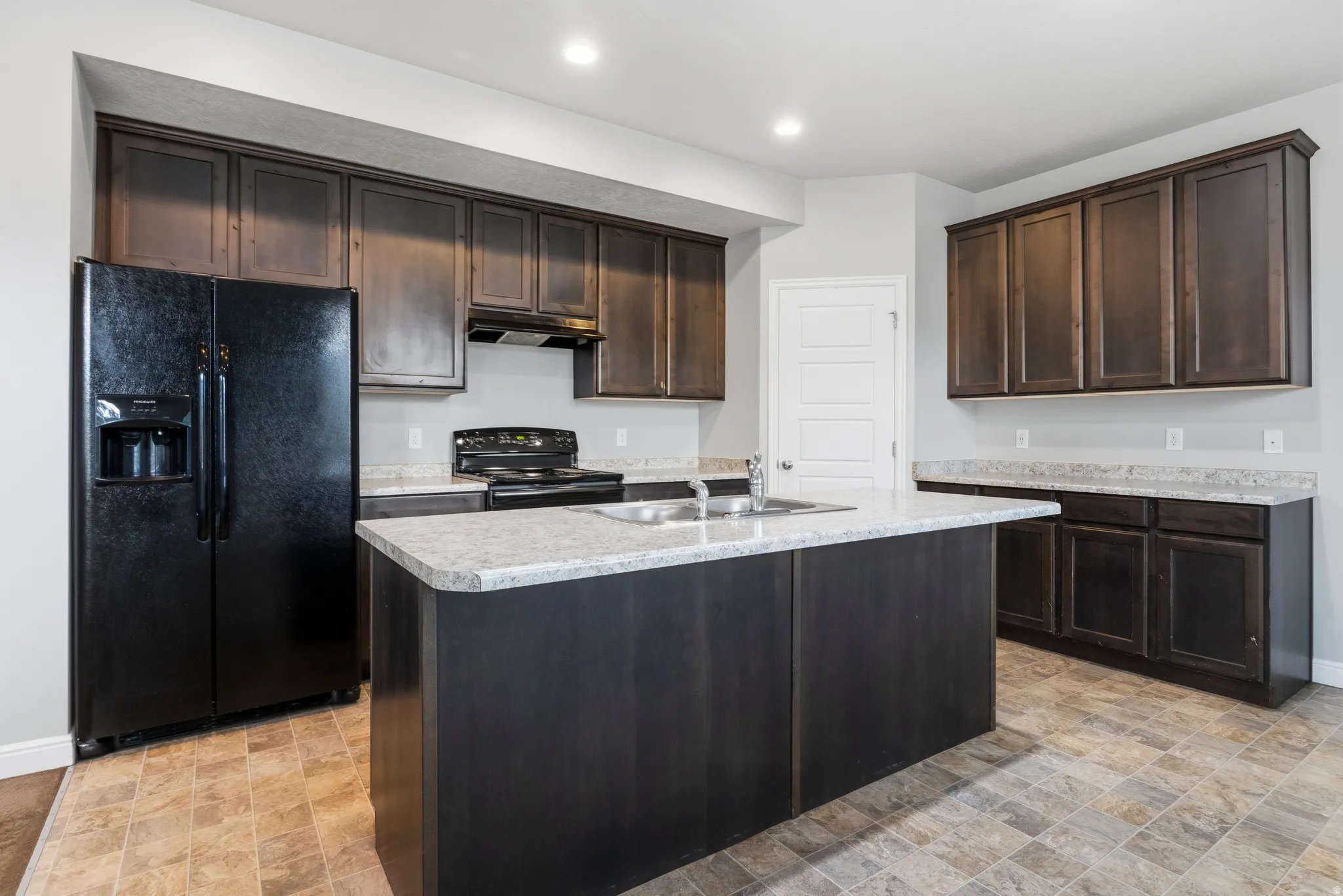 Kitchen featuring dark wood-finish cabinetry, light-toned countertops, a central island with an integrated sink, a black side-by-side refrigerator, and a black range