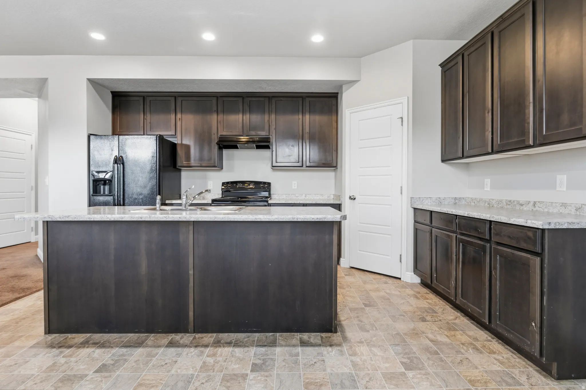 Spacious kitchen featuring an expansive center island with an integrated sink, dark wood-finish cabinetry, light-toned countertops, and a full suite of black appliances