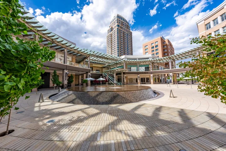 Central courtyard featuring a large circular fountain with stone-finish surfacing, extensive light-toned paver flooring, and overhead architectural trellises
