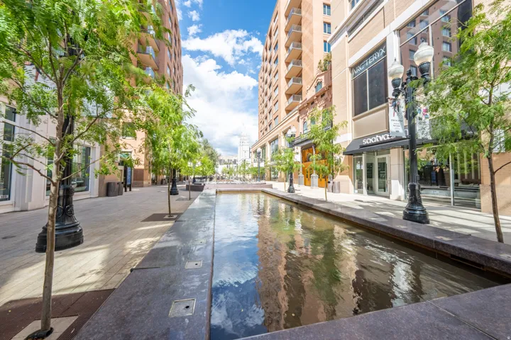 Pedestrian-friendly urban walkway featuring a reflective water feature, mature tree plantings, street lighting, and retail frontage with large display windows