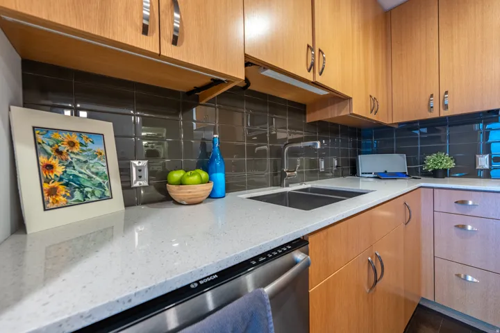 Light wood-finish cabinetry, under-cabinet lighting, stainless steel sink, pull-down faucet, and a grey subway tile backsplash
