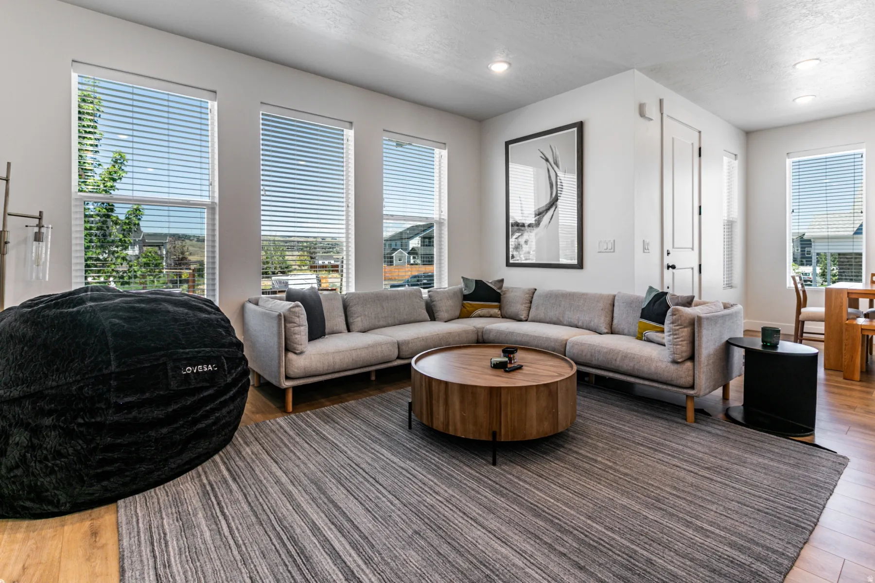 Bright living space featuring multiple large windows with horizontal blinds, wood-finish flooring, recessed lighting, and a neutral color palette