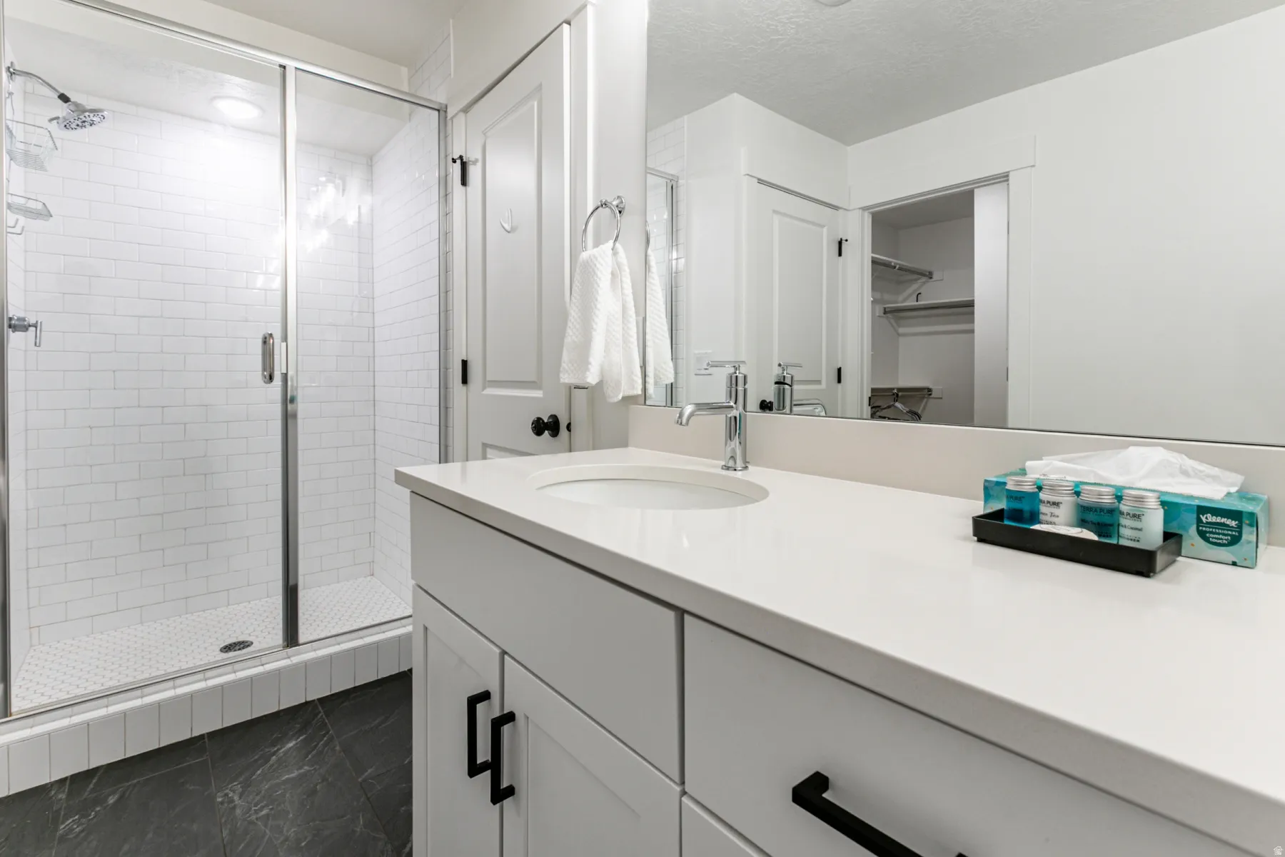 Bathroom featuring a white subway tile shower with a glass enclosure, white vanity with a single basin sink, polished chrome faucet, and dark-toned floor tiling
