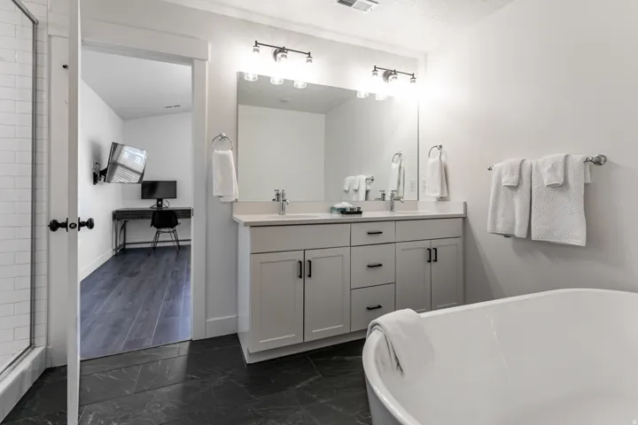 Bathroom featuring a freestanding tub, dark stone-finish flooring, a double vanity with light cabinetry, and a large wall mirror with vanity lighting