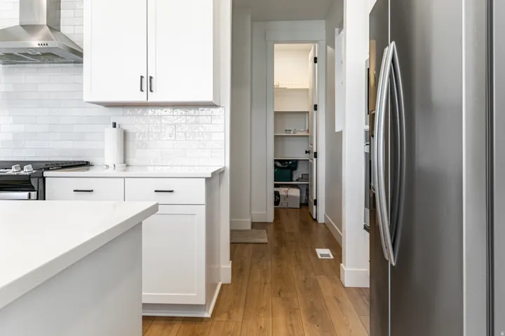 Modern kitchen featuring a stainless steel range hood, white subway tile backsplash, white cabinetry with dark hardware, a stainless steel refrigerator, and wood-finish flooring