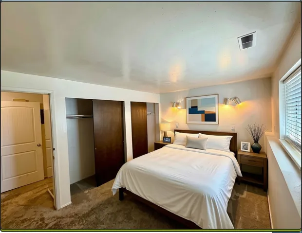 Spacious master bedroom featuring dual wood-panel sliding closet doors, wall-mounted lighting fixtures, a large window with an abundance of light, and neutral-toned wall-to-wall carpeting